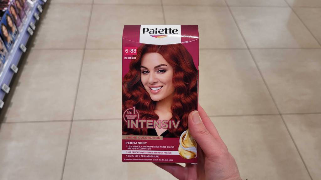 Eine Hand hält eine Packung getesteter roter Haarfarbe vor dem Boden eines Drogeriemarktes.