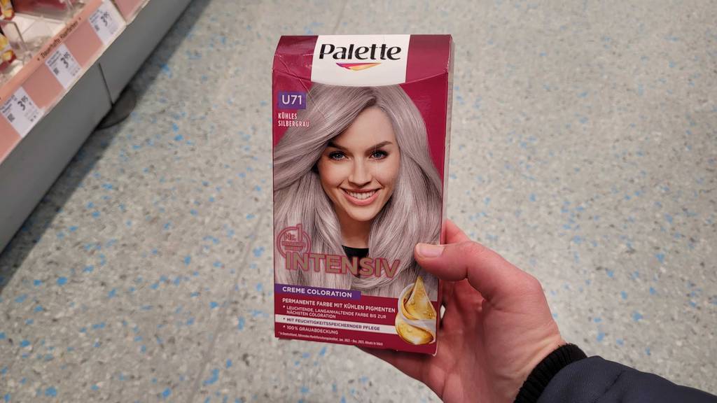 Eine Hand hält eine Packung mit getesteter Silber-Haarfarbe vor dem Boden eines Drogeriemarktes.