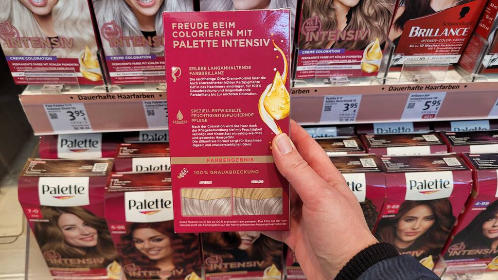 Eine Hand hält eine Packung mit Fokus auf die Produktbeschreibung einer Silber-Haarfarbe im Test vor einem Regal mit Haarpflegemitteln.