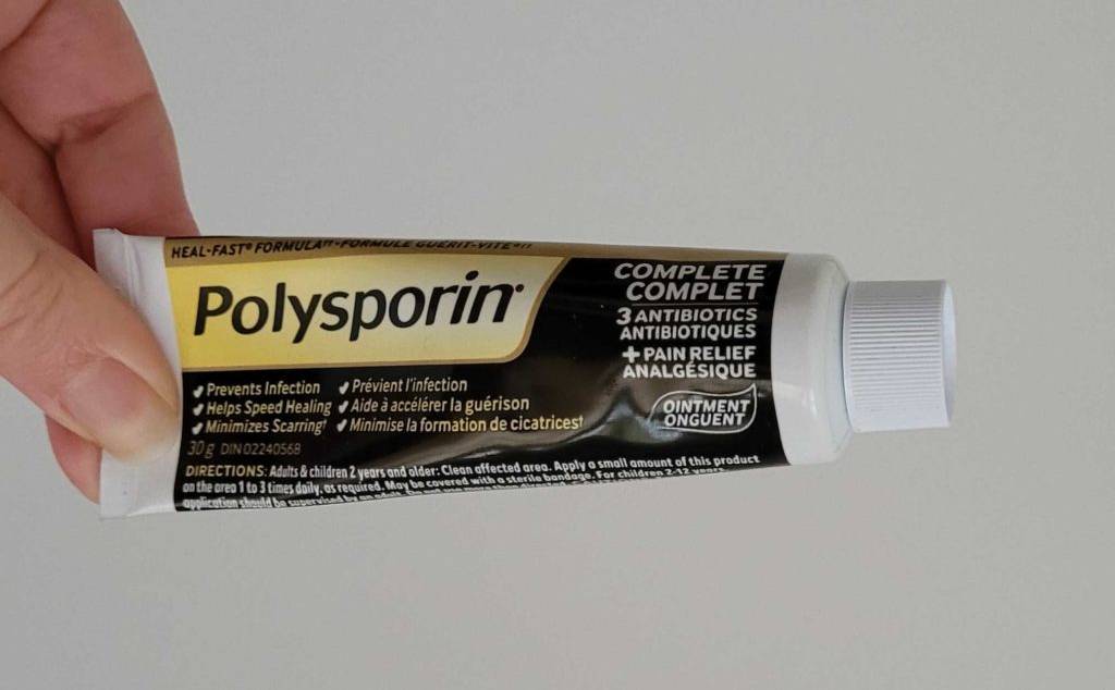 Eine kleine schwarz-weiß-gelbe Tube mit Polysporin Narbengel im Test in einer Hand vor hellem Hintergrund.