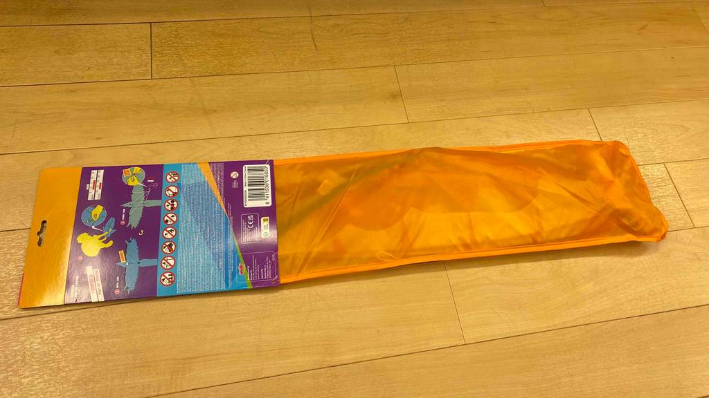 Originalverpackung eines getesteten Kinderdrachens liegt mit der Rückseite nach oben quer auf einem hellen Holzboden.
