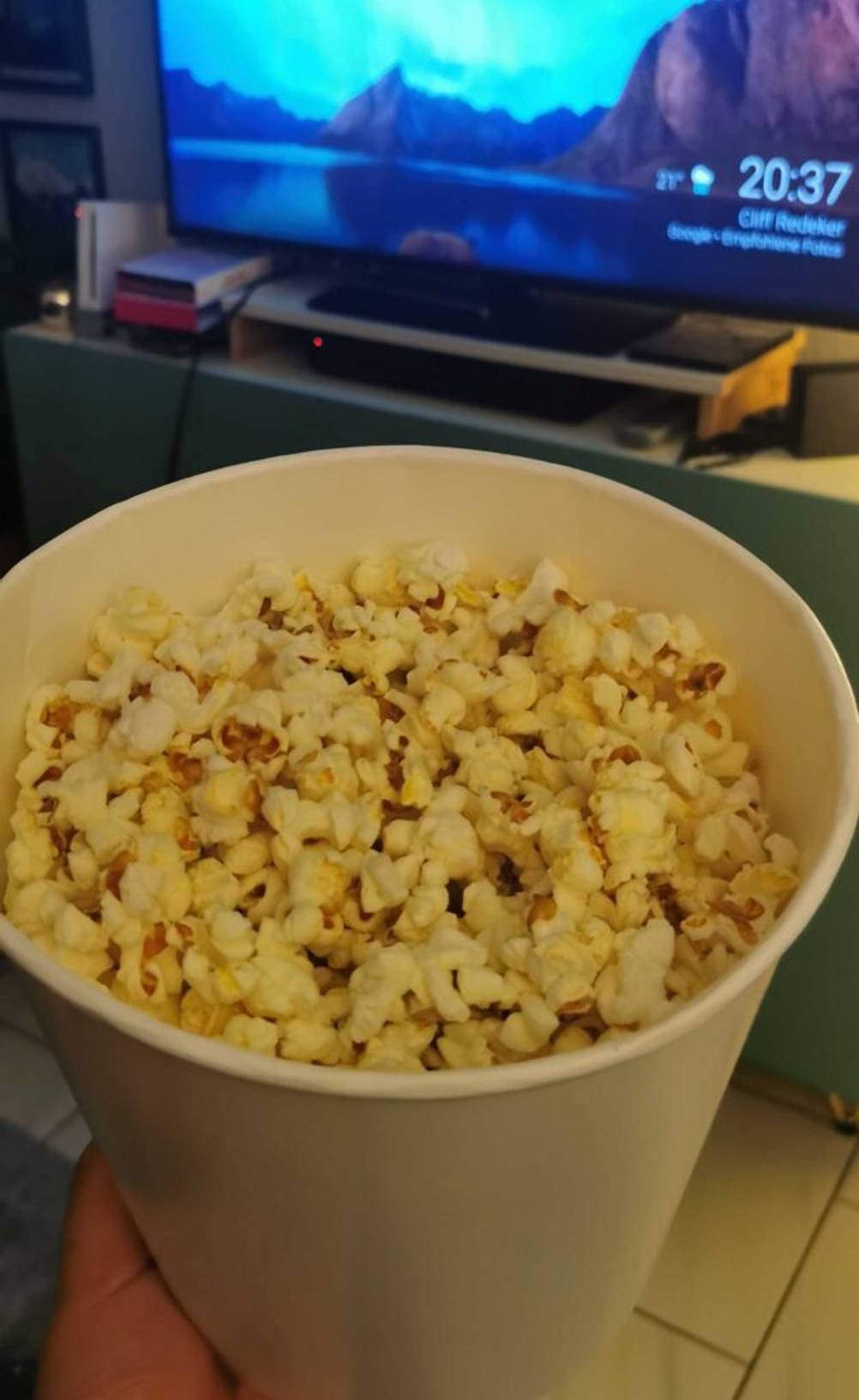 Hand hält Popcornmais im Test in einem weißen Behälter; im Hintergrund befindet sich ein eingeschalteter Fernseher.