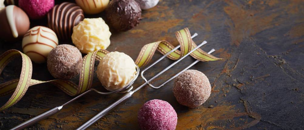 Pralinengabel-Test: Verschiedene runde Pralinen liegen auf einer dunklen Oberfläche. In der Mitte sind eine runde Pralinengabel und eine Pralinengabel mit drei Zinken abgelegt.