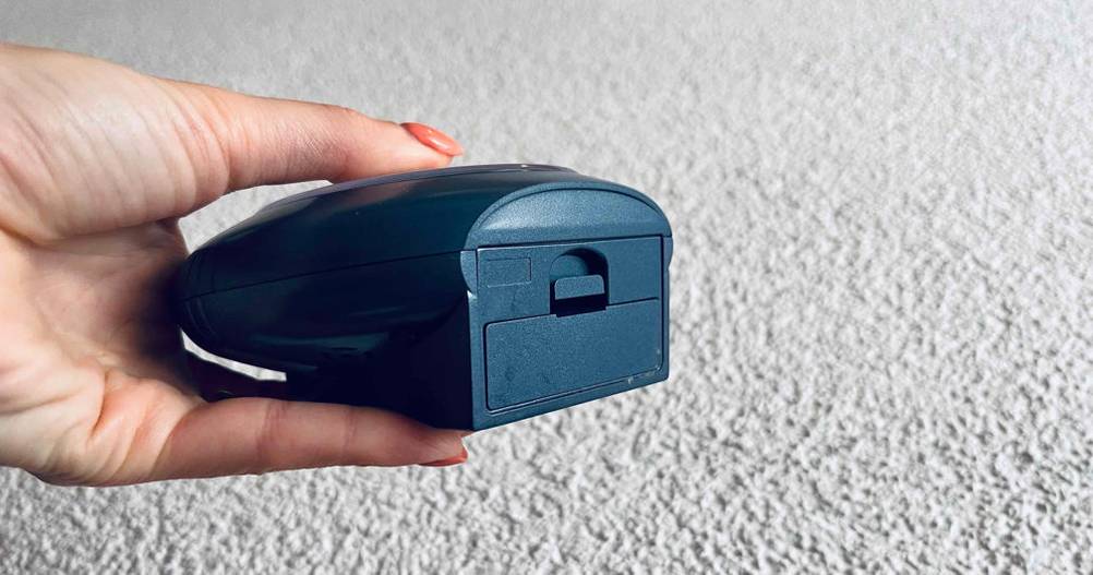 Hand hält einen schwarzen, analogen Funkwecker im Test, mit Fokus auf das geschlossene Batteriefach vor weißer Rauputzwand.