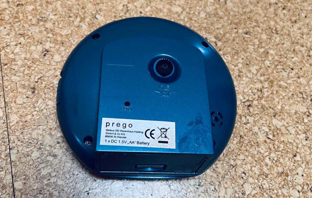 Schwarzer analoger Funkwecker im Test liegt mit der Rückseite inklusive Einstellrad nach oben auf einem hellbraun gesprenkelten Fliesenboden.