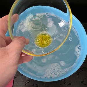 Eine Hand hält eine nasse, durchsichtige Glasschale, in der sich Olivenöl befindet, fest.