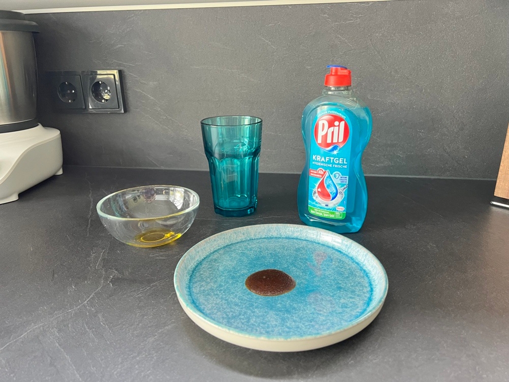 Auf einer Küchenarbeitsplatte stehen ein Teller mit einem eingetrocknetem Fleck Ketchup, eine Schale mit Olivenöl und ein Glas mit eingetrockneter Milch.