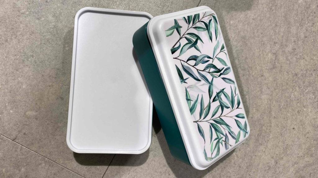 Zwei grün-weiße getestete Lunchbox-Modelle mit floral designtem Deckelbefinden sich auf hellgrauen Fliesen.