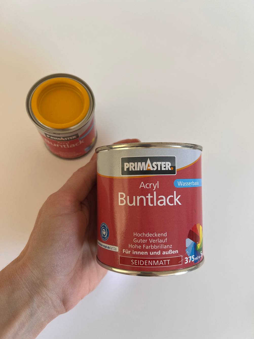 Eine Dose Maleracryl in einer Hand, dahinter eine offene Dose.
