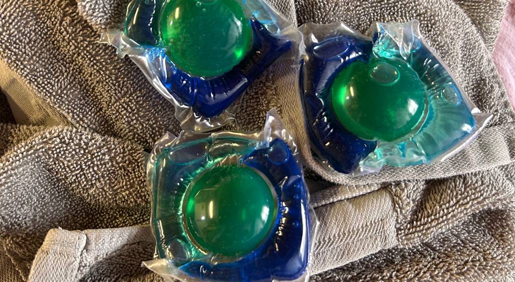 Drei Waschmittel-Pods im Test liegen in wasserlöslicher Kunststoffverpackung auf einem beigefarbenen Handtuch.