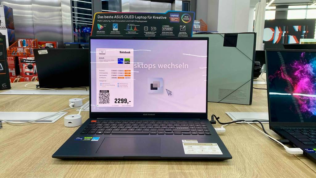 Ansicht von vorn auf ein im Geschäft ausgestelltes schwarzes OLED Notebook ASUS-VivoBook im Test, dessen glänzender Bildschirm eingeschaltet ist.