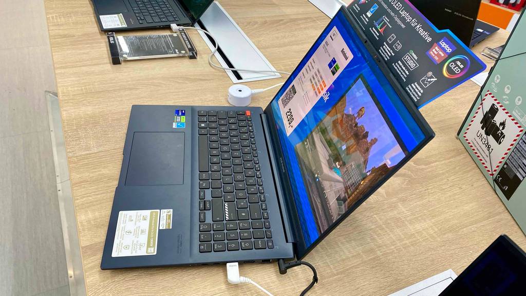Im Test: ein ASUS-VivoBook in Schwarz befindet sich eingeschaltet mit glänzendem Bildschirm neben weiteren Laptops auf einem Holztisch im Geschäft.