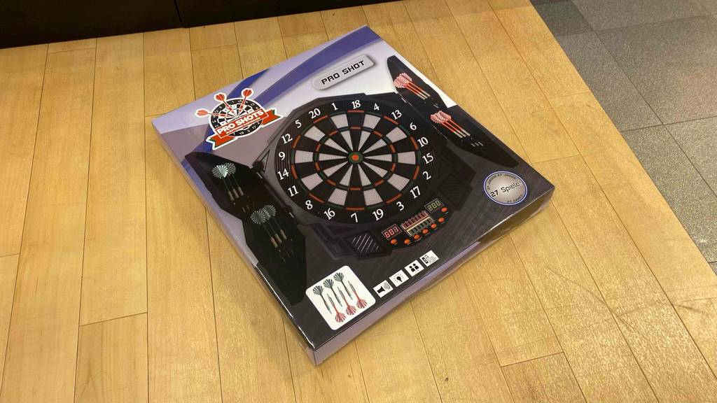 Originalverpackung einer getesteten Dartscheibe liegt schräg auf einem Holzboden.