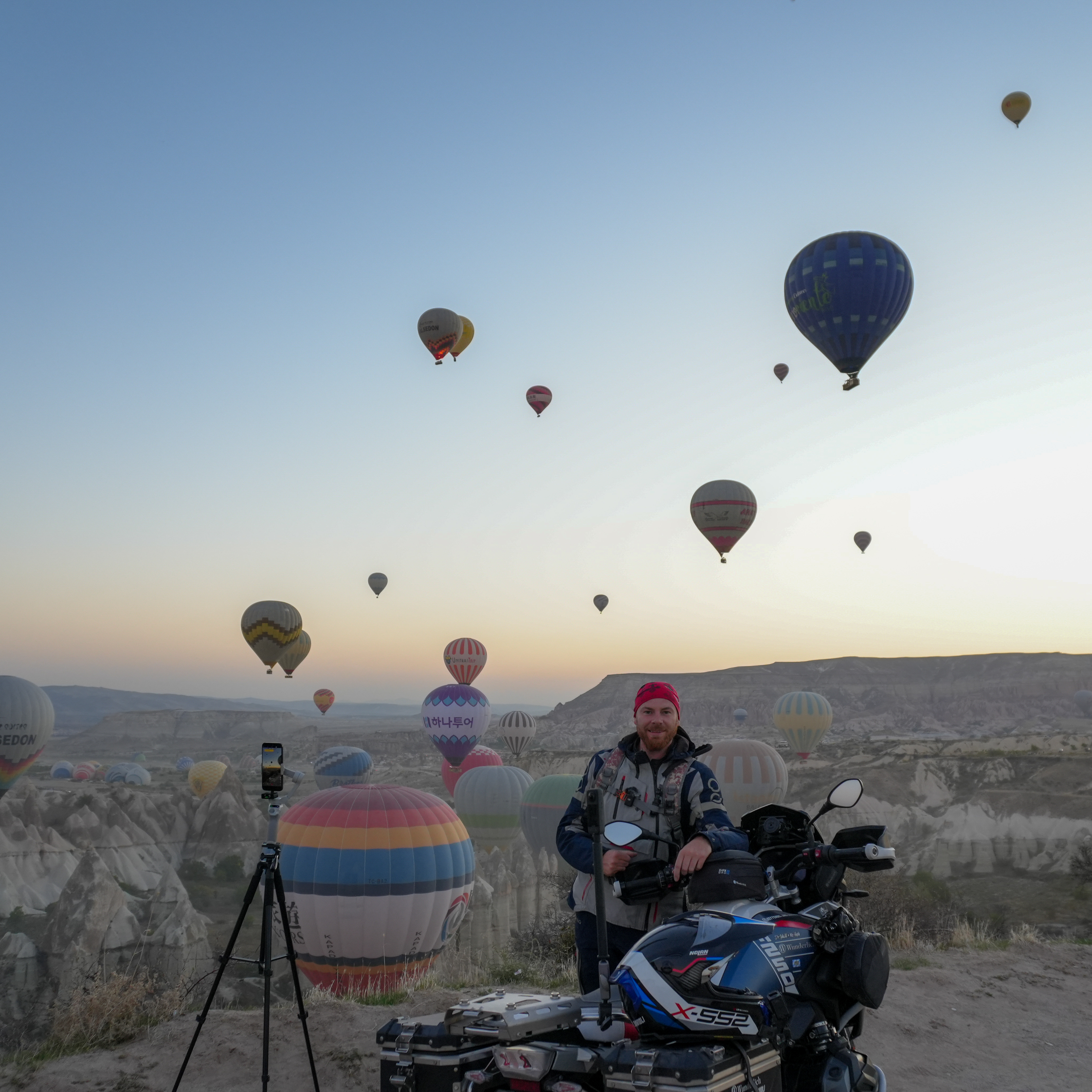 VGL-Motorradexperte Meli Schönbrodt mit seinem Motorrad in einer Steppenlandschaft umgeben von aufsteigenden Heissluftballons.
