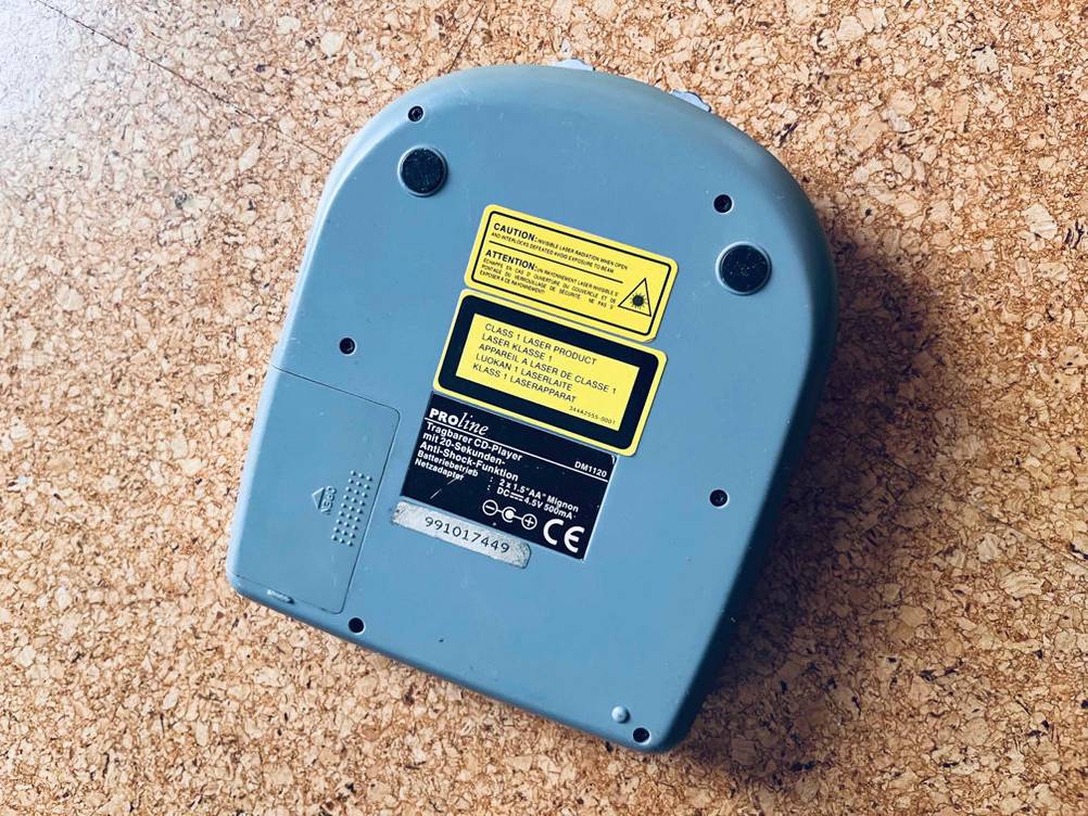 Hellblauer tragbarer CD-Player im Test befindet sich mit der Standfläche nach oben inklusive Sicht auf das Batteriefach und gelben Anwendungswarnungen auf einem braun gesprenkelten Boden.