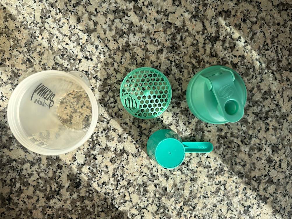 Eiweiß-Shaker im Test: Ein Set mit Becher, Einsatz, Deckel und Dosierlöffel, das auf einer Küchenarbeitsplatte steht, von oben betrachtet.