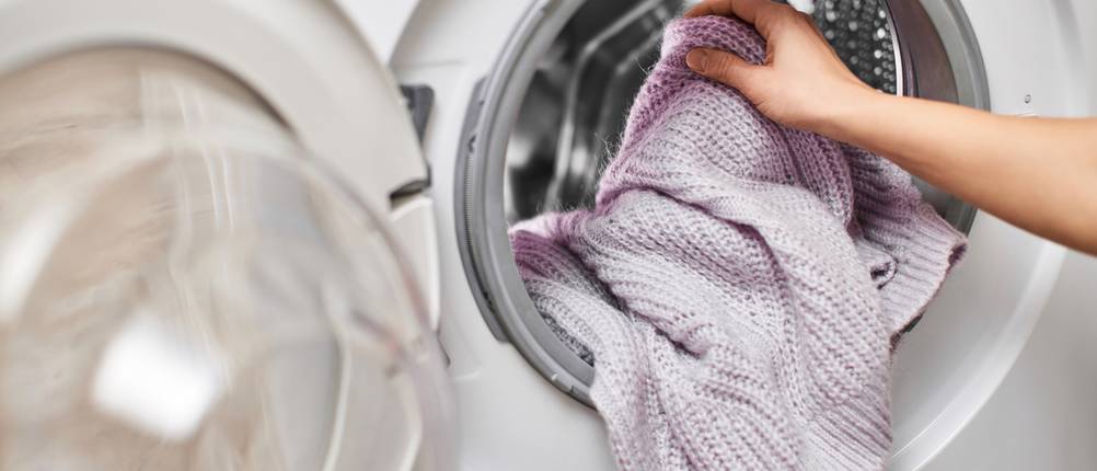 Energiesparende-Waschmaschinen-Test: Fliederfarbener Pullover wird von einer Hand in eine Waschmaschine gelegt.