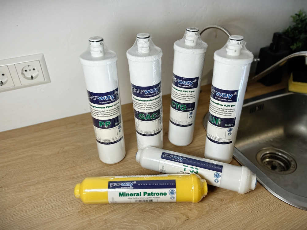 Sechs Purway-Ersatzfilter unterschiedlicher Typen liegen nebeneinander auf einer Küchenarbeitsplatte. Im Hintergrund ist eine Spüle mit Wasserhahn zu sehen.