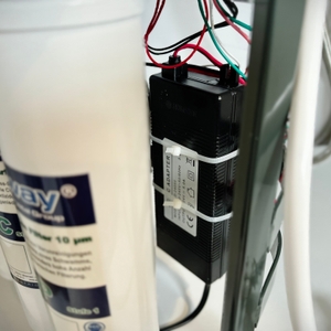 Ein schwarzes Netzteil ist mit Kabelbindern an einem Metallrahmen befestigt. Mehrere farbige Kabel führen in die Anlage.