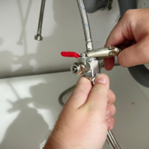 Zwei Hände montieren ein T-Stück mit Absperrhahn an eine metallene Wasserleitung unter der Spüle. Der rote Hebel steht offen.