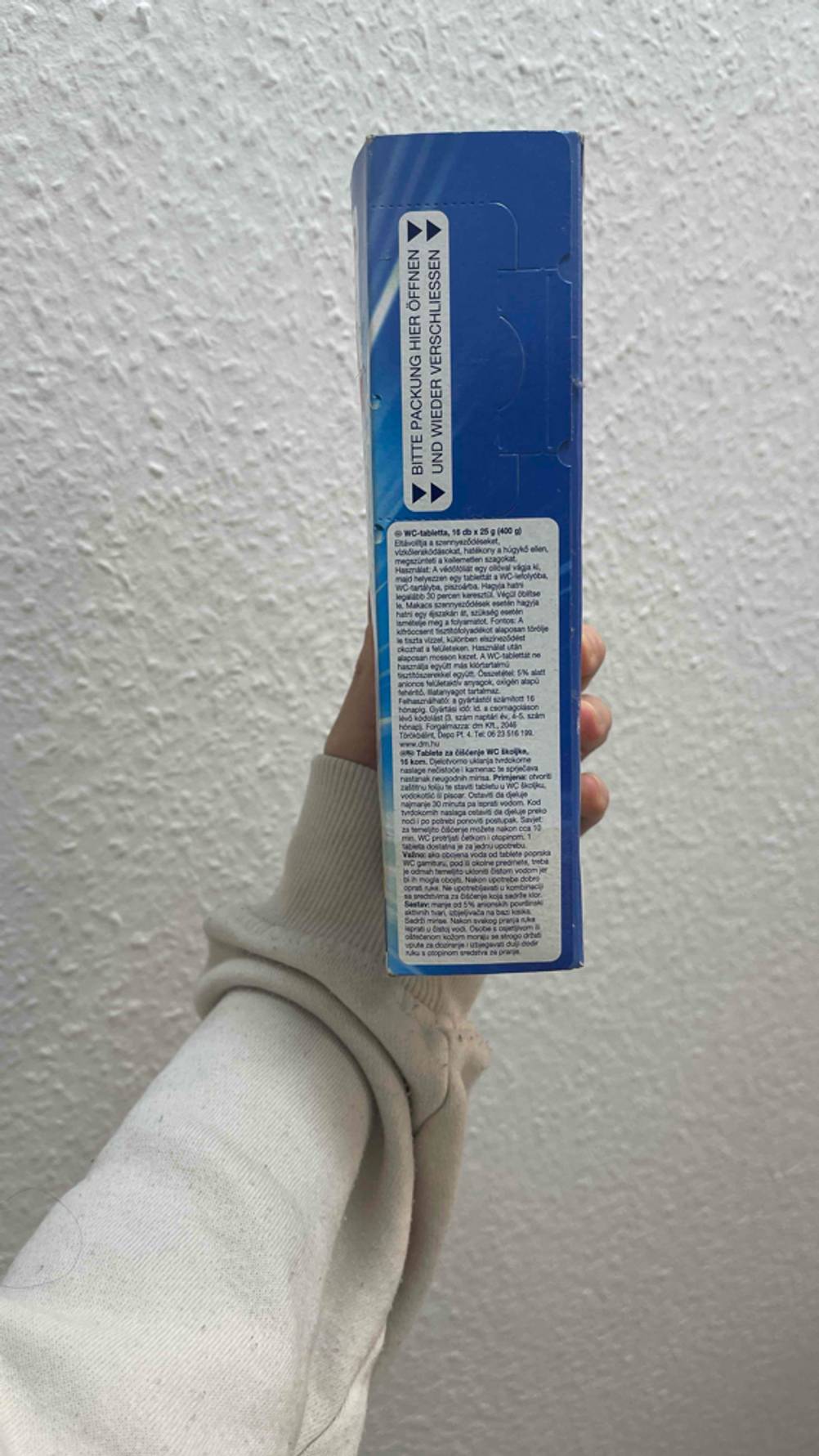 Eine Hand hält die Verpackung der getesteten Putzmittel-Tabs seitlich vor eine Wand.