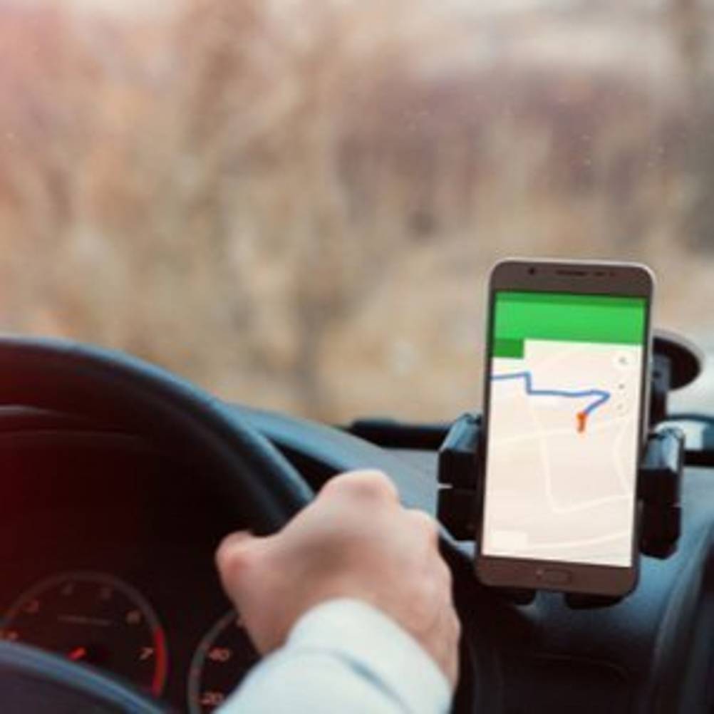 Mann, der in einem Auto fährt und dabei auf das Smartphone schaut, auf dem eine Navigationsapp läuft und das mithilfe eines Qi-Ladegerätes mit Saugnapf an der Windschutzscheibe befestigt ist