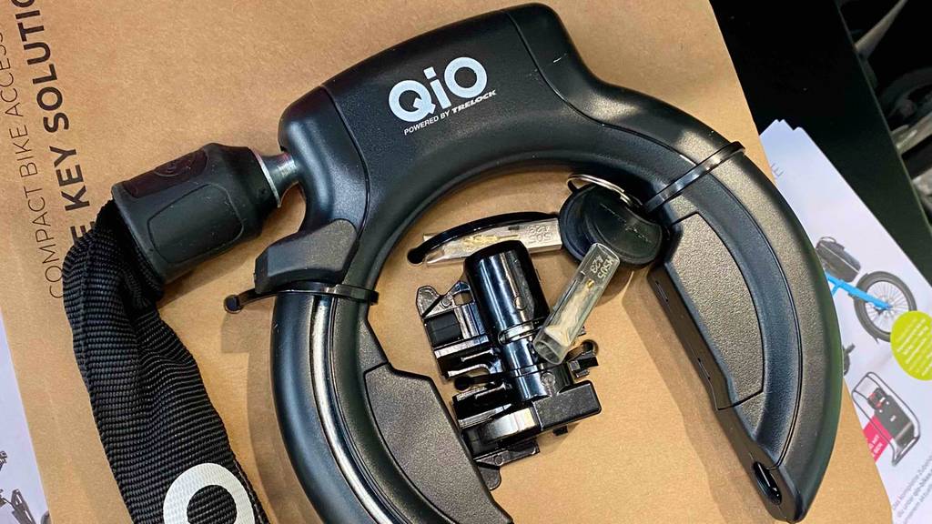 Auf einem braunen Karton ist ein schwarzes Fahrrad-Rahmenschloss mit Kette im Test befestigt. Das Produkt ist von der Marke Qio und mit mehreren Schlüsseln ausgestattet.