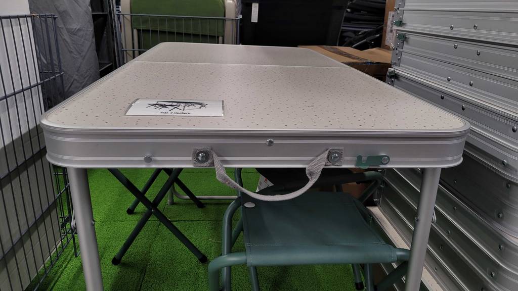 Campingtisch-Set-Test: Ansicht von vorn auf einen silberfarbenen, aufgebauten Campingtisch aus Leichtmetall, darunter stehen grüne Klapphocker.