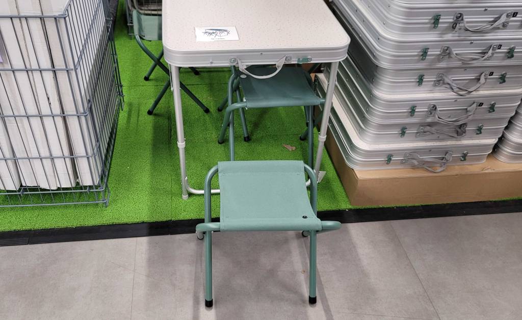 Ansicht von schräg oben auf den grünen Klapphocker und den aufgebauten Campingtischkoffer eines silberfarbenen Campingtisch-Sets im Test, das in einem Verkaufsbereich steht.