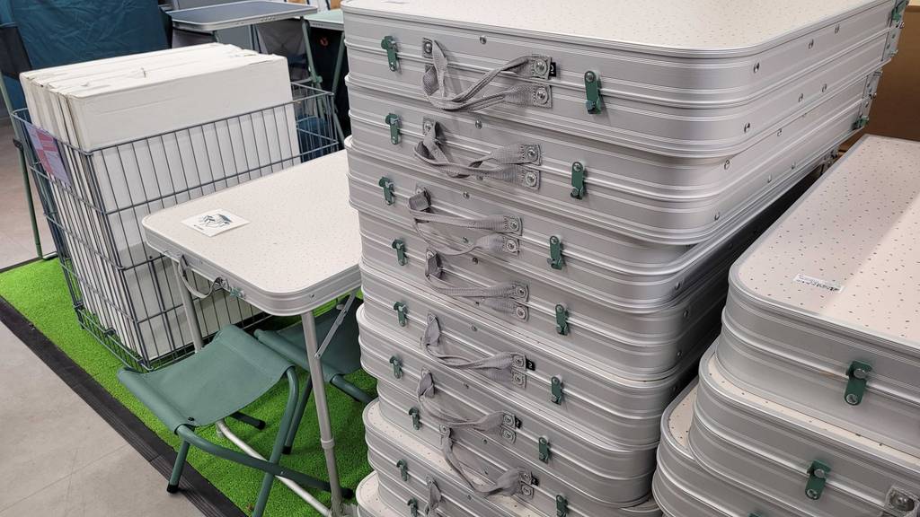 Mehrere Campingtischkoffer befinden sich auf einem Stapel neben einem ausgestellten, getesteten Campingtisch-Set inklusive Hocker im Geschäft.