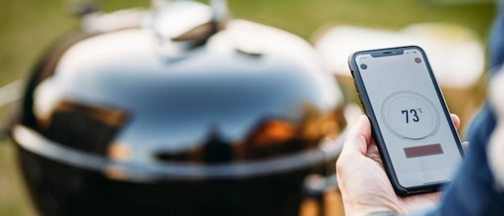 Digitale Räucherthermometer im Test: Person liest Temperatur auf einer App auf dem Smartphone ab, ein Grill steht im Hintergrund.