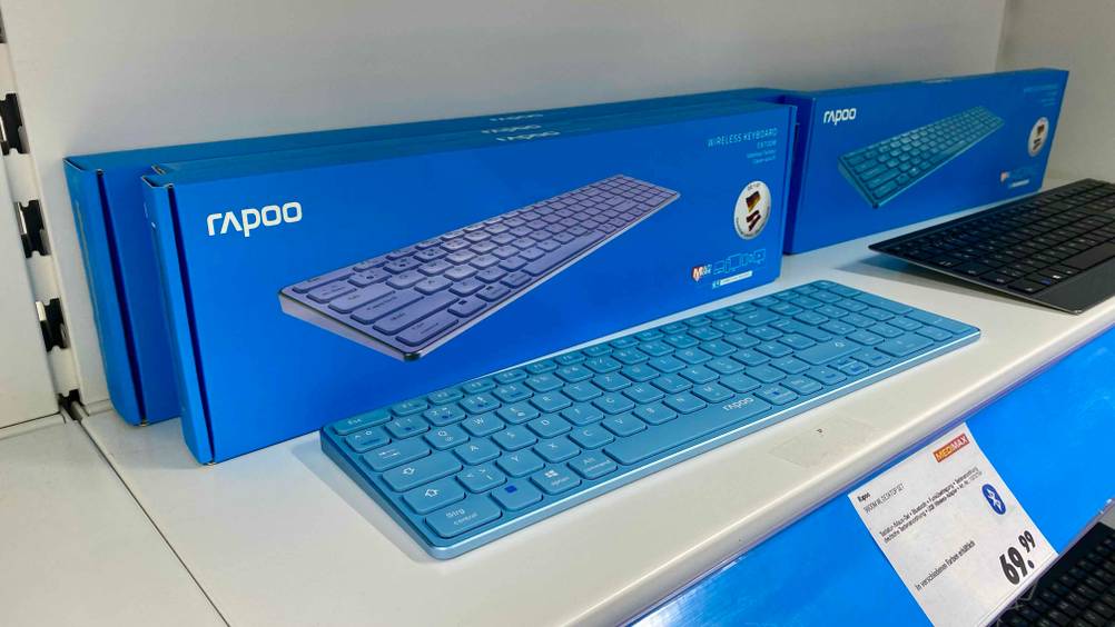 Bluetooth-Tastatur geteste: Ein Gerät auf einer hellen Fläche im Vordergrund Preisangaben im Hintergrund Verpackungen und ein anderes Gerät.
