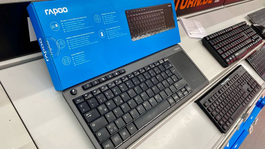Eine schwarze Tastatur mit Touchpad im Test steht vor ihrer blauen Verpackung mit rückseitiger Produktbeschreibung, auf einem weißen Verkaufsregal mit weiteren Tastaturen im schwarzen Design.