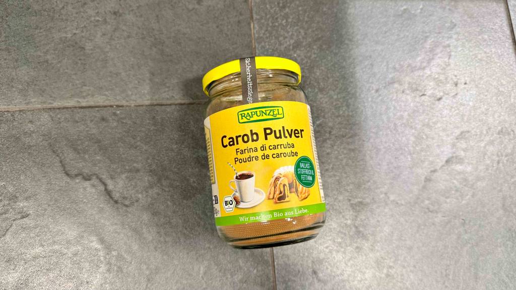 Carobpulver im Test: Ein wiederverschließbares Glas mit braun-gelbem Pulver liegt auf einer grauen Oberfläche.