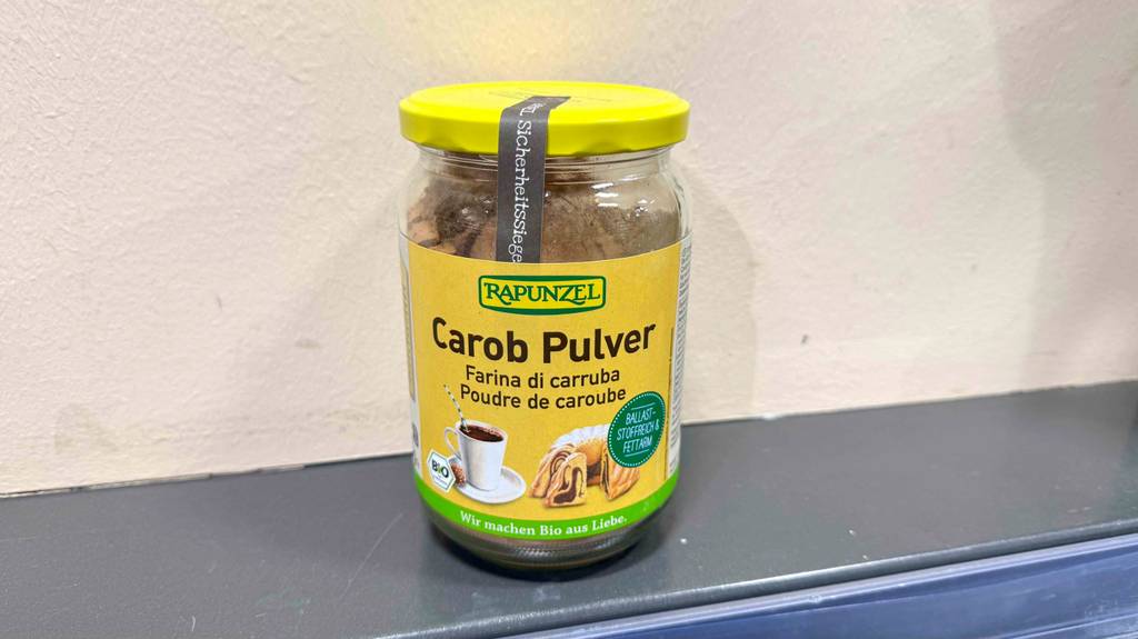 Ein Glas mit getestetem Carobpulver steht vor einer weißen Wand.