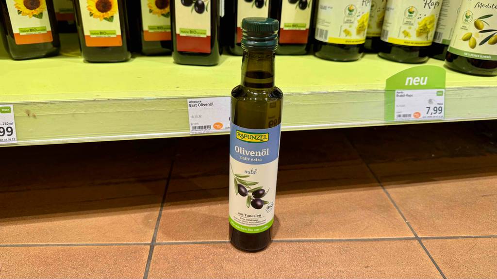 Eine Flasche getestetes mildes Olivenöl steht auf dem Boden vor einem Regal.
