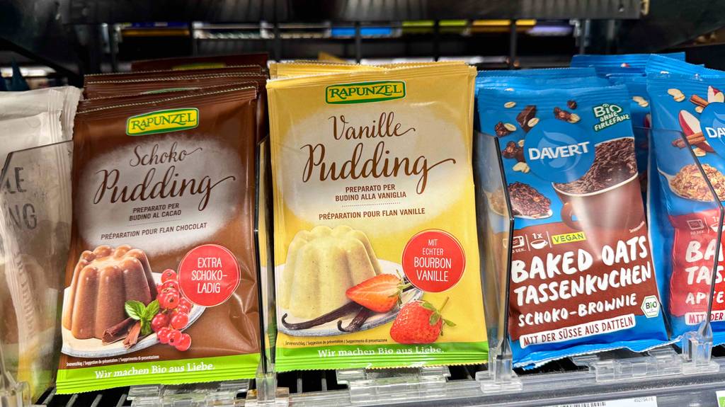 Vanillepuddingpulver getestet: Eine Tüte mit Pulver sowie echter Bourbon-Vanille steht auf einem Regal zwischen weiteren Puddingsorten.