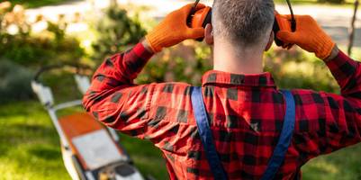 Ein Arbeiter setzt neben einem Rasenmäher Gehörschutz auf.