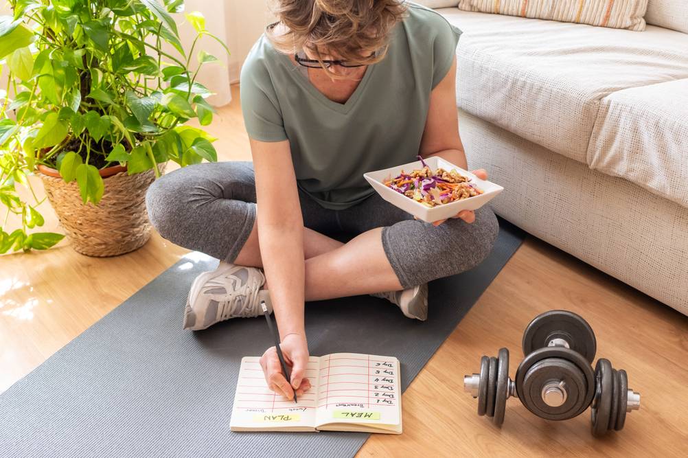 Eine Frau sitzt auf einer Yogamatte. In einer Hand hält sie eine Schüssel mit Salat, während sie gleichzeitig in ein Tagebuch schreibt