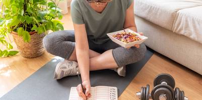 Eine Frau sitzt auf einer Yogamatte. In einer Hand hält sie eine Schüssel mit Salat, während sie gleichzeitig in ein Tagebuch schreibt