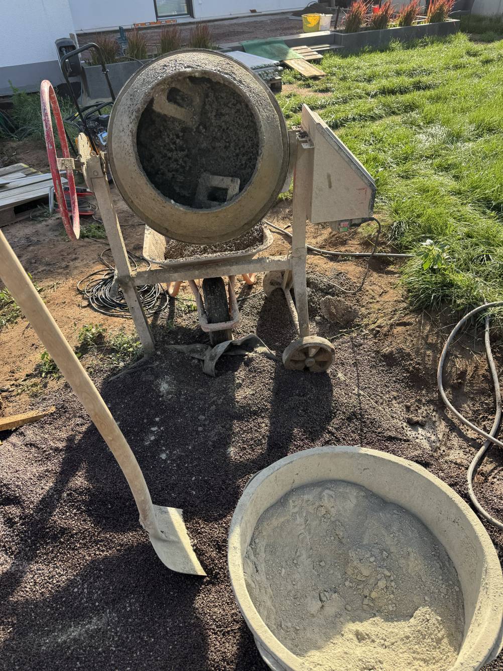 Drainagebeton wird in einem Betonmischer in einem Garten angemischt. Vor dem Mischer steht ein Eimer mit Trasszement.