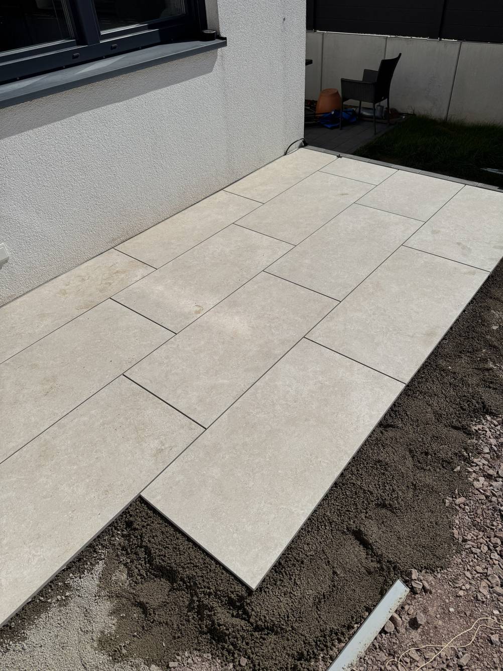 Eine Baustelle einer Terrasse, bei der große rechteckige Keramik-Terrassenplatten auf Drainagemörtel vor einem Haus verlegt werden.