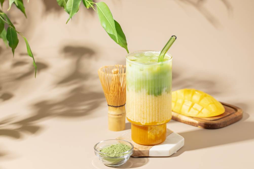 Mango Matcha Getränk in einem Glas mit Strohhalm steht auf einem Tisch. Im Hintergrund liegt eine Scheibe Mango, vorn ist Matcha Pulver in einem kleinen Glasschälchen. Ein Matscha-Teepinsel steht hinter dem Glas.