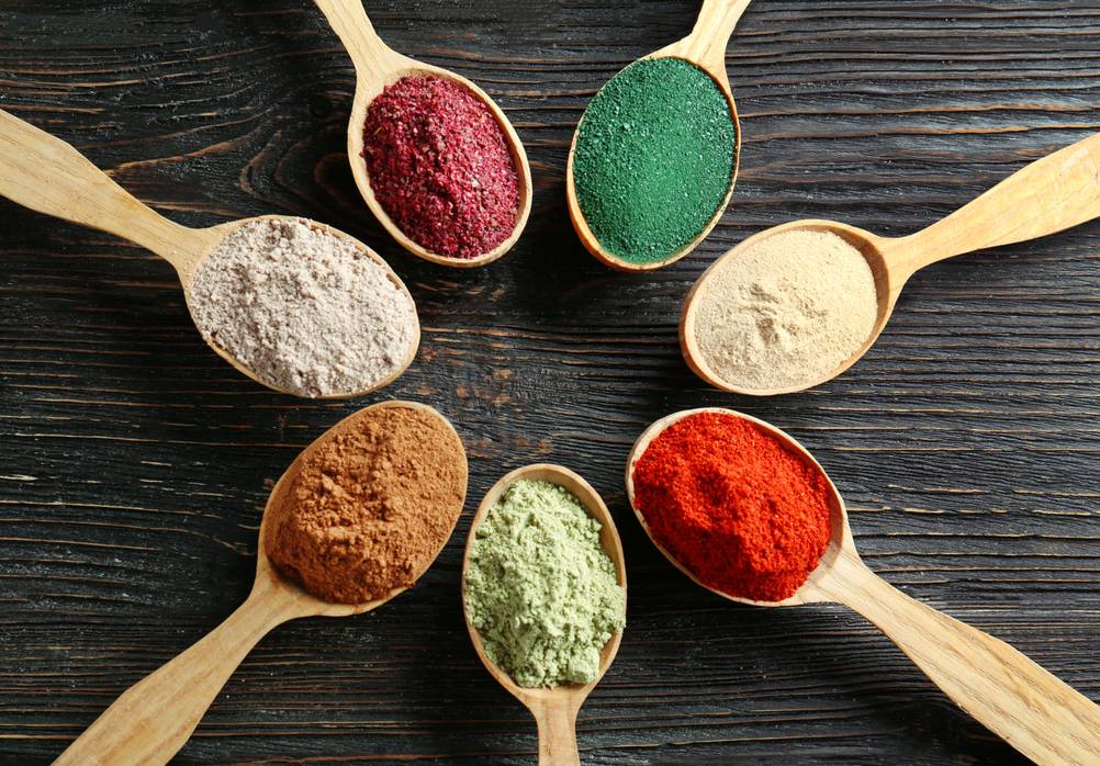 7 verschiedene Superfood-Pulver liegen in Holzlöffeln in einem Kreis.