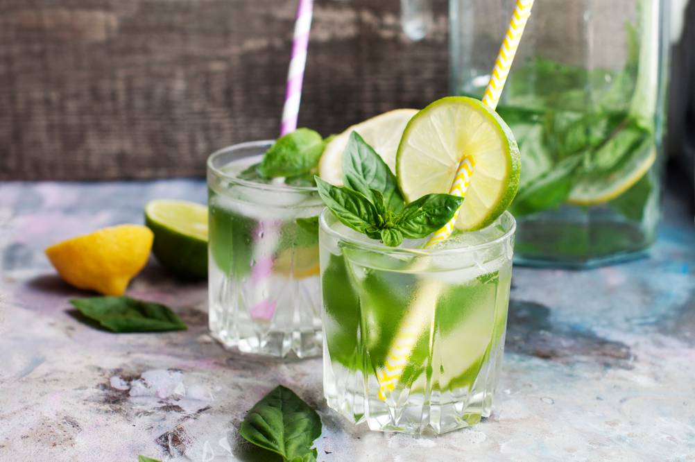 Zwei Gläser Limonade mit Limettenscheiben, Minz- und Basilikumblätter stehen auf einem Tisch.