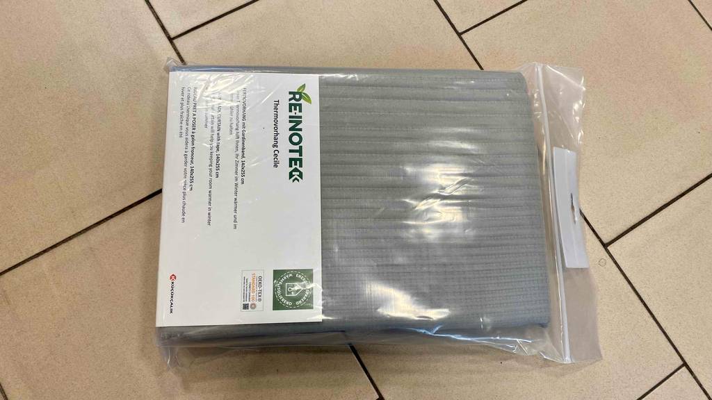 Thermovorhang-Test: hellgrauer Stoffvorhang von Re-inote zusammengefaltet in einer durchsichtigem Kunststoffverpackung auf einem Fliesenboden.