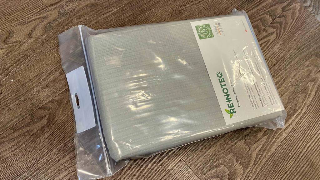 Thermovorhang getestet: Ein hellgrauer Stoffvorhang von Re-inote befindet sich zusammengefaltet in einer durchsichtigem Kunststoffverpackung auf einem Holzboden.