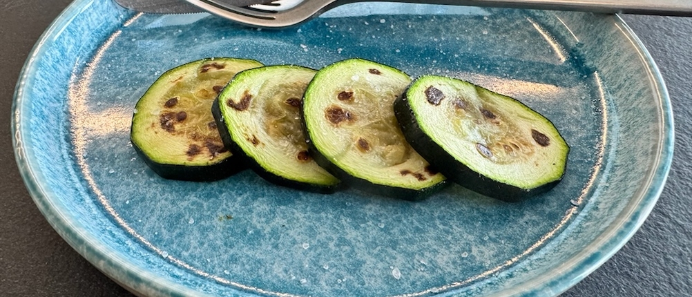 Vier Zucchinischeiben liegen mit Messer und Gabel im Hintergrund auf einem blauen Teller