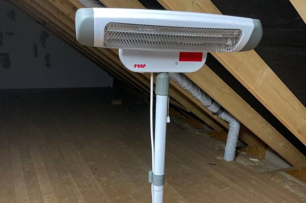 Weißer Reer Heizstrahler im Test mit Paneel steht inklusive Teleskop-Ständer auf einem Dachboden aus Holz.