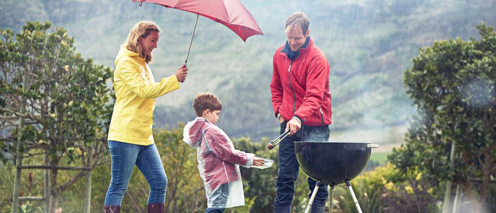 Mutter, Vater und Kind grillen bei Regen im Garten.